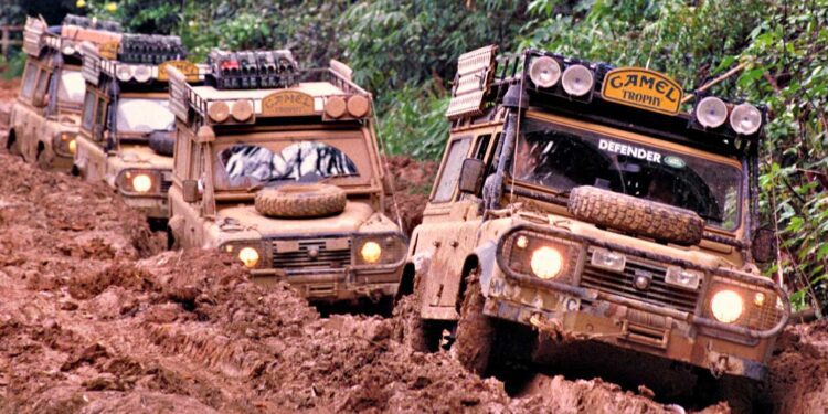 Three mud-covered Land Rover Defenders, true historic expedition icons, drive in a line through a narrow, muddy off-road track surrounded by dense green vegetation.