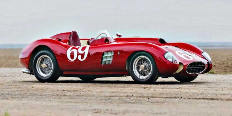 A classic red Ferrari 375 Plus race car with the number 69 on the side and hood is parked on a sandy surface under a cloudy sky.