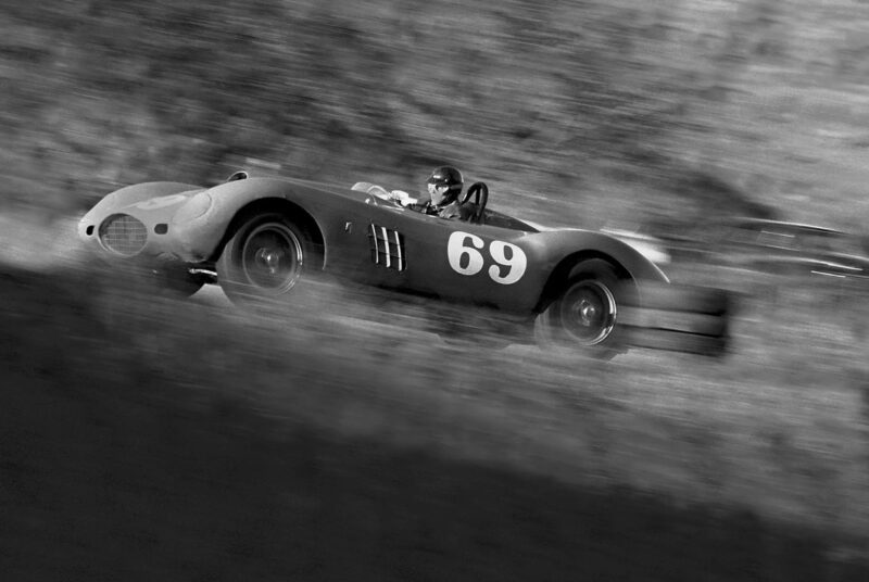 A vintage Shelby race car with the number 69, driven by a helmeted driver, speeds along a track, captured in motion blur in a timeless black and white photo.