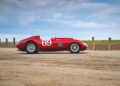 A classic red Ferrari 375 Plus race car with the number 69 on its side is parked outdoors on a dirt surface, with a cloudy sky and wooden posts in the background—a nod to Shelby and Miles’ legendary racing era.