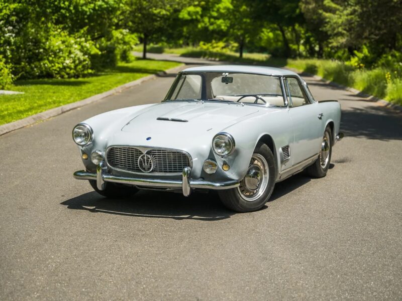 A classic silver Maserati coupe, exuding Italian luxury from the 1950s or 1960s, is parked on a paved road surrounded by greenery and trees on a sunny day.