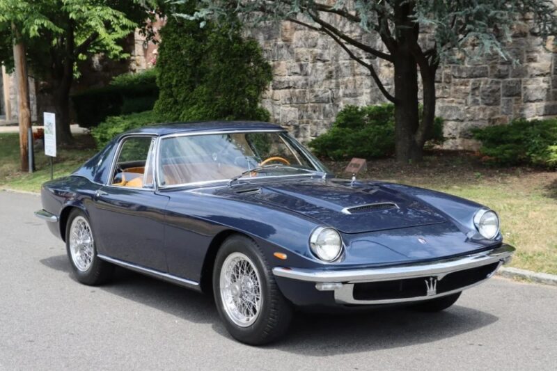 A classic blue Maserati sports car, a true icon of Italian luxury, is parked on a street near stone walls and greenery.
