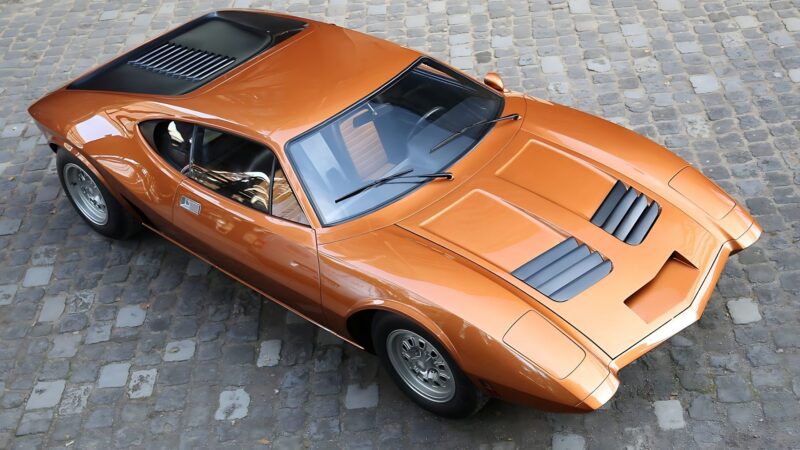A bronze-colored vintage sports car, one of the 10 Unsung Mid-Engine American Concepts That Pushed the Envelope Of Performance, is parked on a cobblestone surface and viewed from above at an angle.