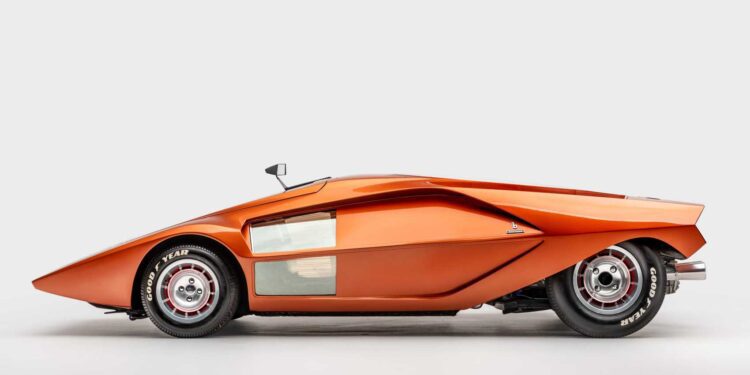 Side view of an orange wedge-shaped concept car with a low, angular profile, small side mirror, visible Goodyear tires, and silver panel detailing, as seen in the exhibit at the Petersen Museum against a plain background.