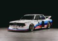 A vintage BMW race car with number 2, featuring a white body, blue and red stripes, and sponsor logos, is parked indoors on a gray concrete floor during Monterey Car Week 2025.