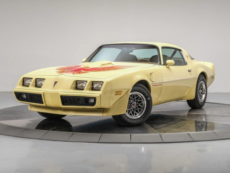 A yellow Pontiac Trans Am sports car with a firebird decal on the hood, parked indoors on a circular platform—a stunning example of Classic Muscle Cars for sale.