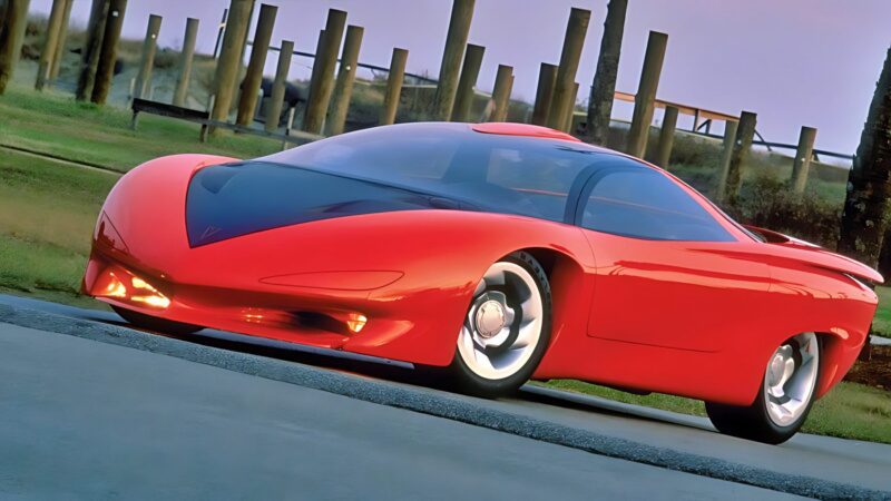 A bright red, futuristic sports car with a sleek design—resembling one of the 10 Unsung Mid-Engine American Concepts That Pushed the Envelope Of Performance—is parked on a concrete surface near grass and wooden posts.