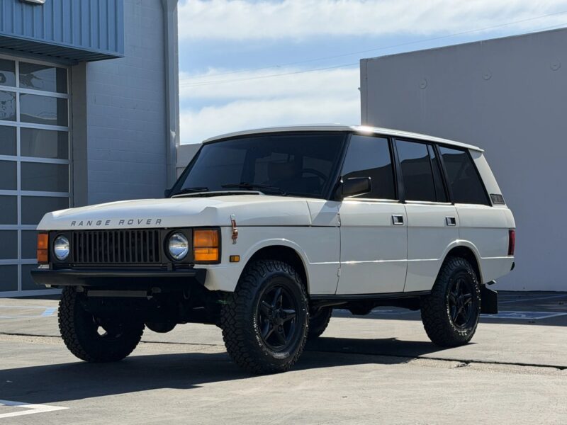 A white classic Range Rover luxury SUV with black wheels is parked in an outdoor lot near a gray industrial building.