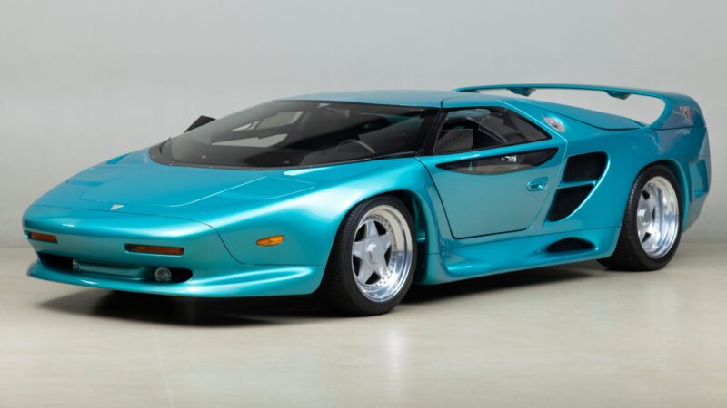 A teal-colored, low-profile sports car with aerodynamic styling, large rear wing, and wide tires—reminiscent of the 10 Unsung Mid-Engine American Concepts That Pushed the Envelope Of Performance—is parked on a light-colored floor against a plain backdrop.