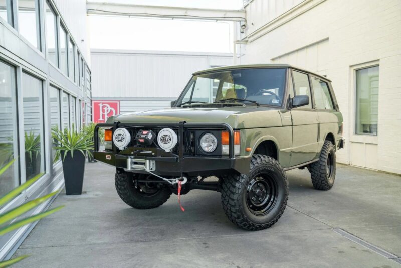 A vintage green Range Rover with off-road tires and a winch is parked in an outdoor area between buildings, showcasing one of the best manual transmission cars for sale.