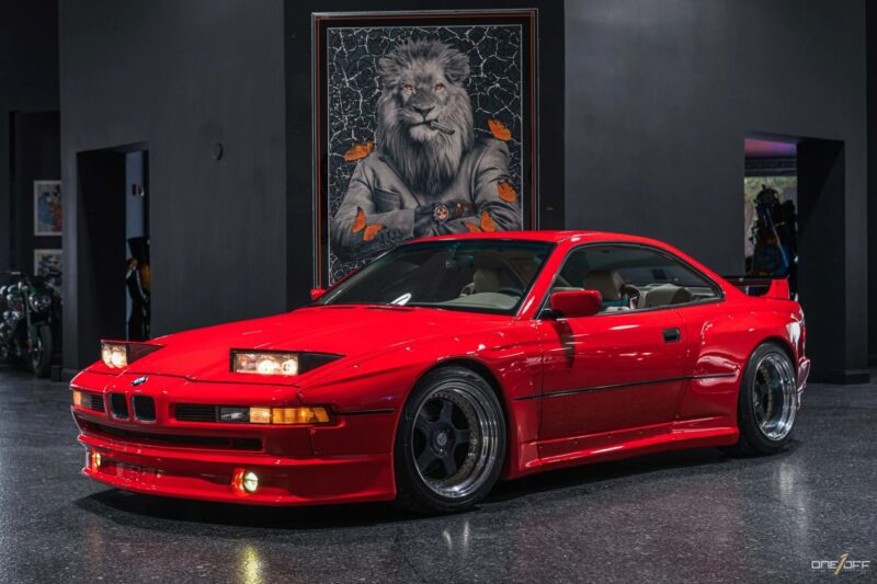 A red BMW coupe with pop-up headlights, a true icon for fans of 90s cars for sale, is parked indoors on a shiny floor. Behind the car is a large framed artwork of a lion wearing a suit and watch.