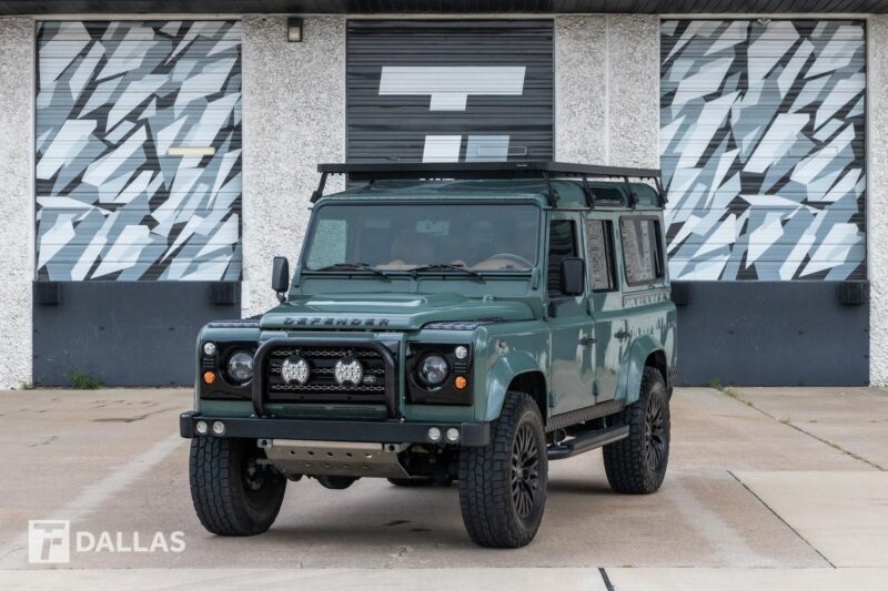 A green Land Rover Defender with a roof rack, echoing nostalgia, is parked in front of a building with patterned garage doors.