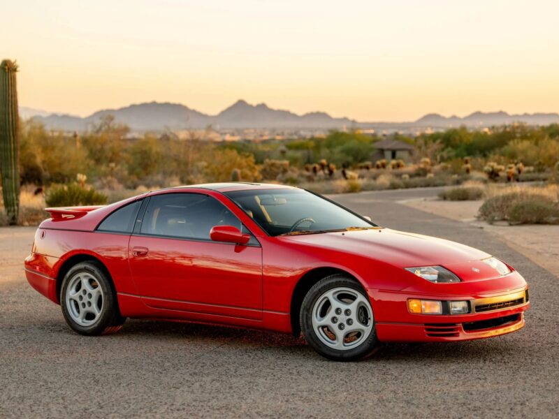 A red two-door sports car is parked on a paved road in a desert landscape with mountains visible in the background at sunset, evoking nostalgia for classic 90s cars for sale and the history they represent.