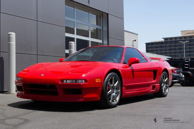 A red Acura NSX, one of the iconic 90s cars for sale, is parked outside on a sunny day near a modern building, with other vehicles visible in the background.