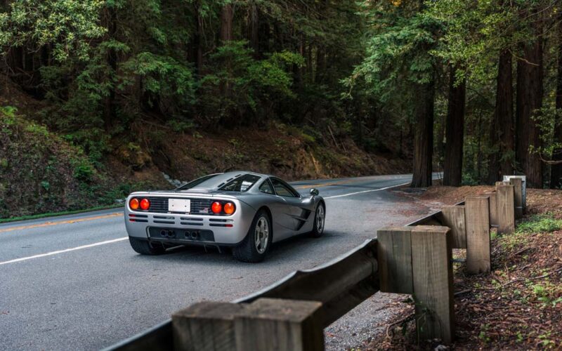 A silver sports car, reminiscent of a McLaren F1, drives along a winding road through a forested area with tall trees and a wooden guardrail—perfect for an RM Sotheby’s Monterey Auction showcase.