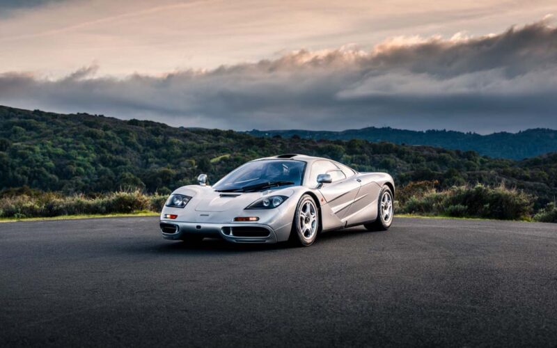 A silver McLaren F1 sports car is parked on an empty paved area, with green hills and a cloudy sky in the background—a stunning sight ahead of the RM Sotheby's Monterey Auction.