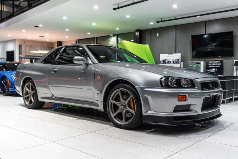 A silver Nissan Skyline GT-R R34 sports car is displayed indoors on a glossy white floor, its showroom lighting and other vehicles evoking nostalgia for 90s cars and the legendary history of this iconic model.