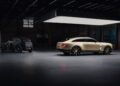 A vintage black car and Bentley’s first-ever EV, a modern beige electric vehicle concept, are parked indoors under a large rectangular ceiling light in a spacious, industrial-style room.