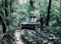 A group of people use ropes to pull a mud-covered vehicle, reminiscent of the Land Rover Classic to Showcase 10 Historic Expedition Icons at 2025 Pebble Beach Concours d'Elegance, stuck in a narrow, waterlogged trail through dense jungle vegetation.