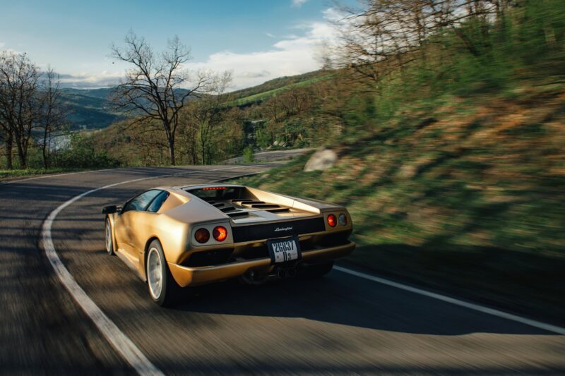 A gold Lamborghini sports car is driving along a winding road surrounded by greenery and trees in Emilia-Romagna under a partly cloudy sky.
