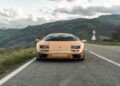 A gold sports car, reminiscent of vintage Lamborghinis, is parked on an empty road in Emilia-Romagna with hills and cloudy skies in the background.