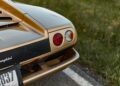 Close-up of the rear left tail lights and bumper of a gold Lamborghini, reminiscent of vintage Lamborghinis, parked on the side of a road next to grass in Emilia-Romagna.