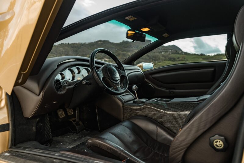 The interior of a sports car with black leather seats and a manual gear shift, reminiscent of Vintage Lamborghinis, offers a stunning view of Emilia-Romagna’s green hills through the windows.