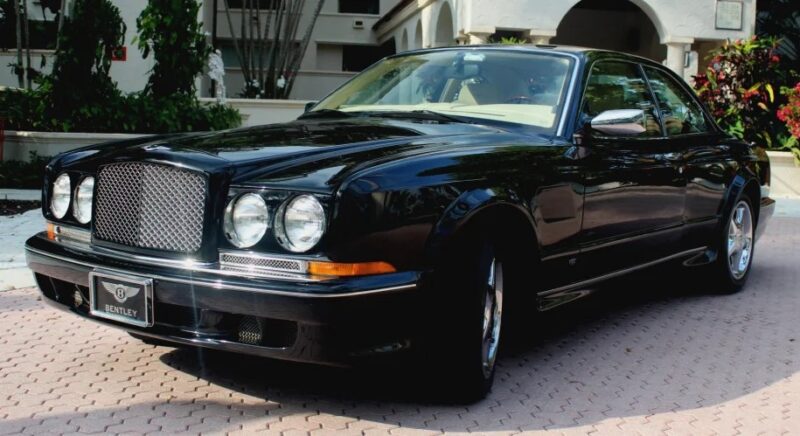 A black Bentley coupe, an exquisite luxury car, is parked on a brick driveway in front of a building with arches and lush landscaping.