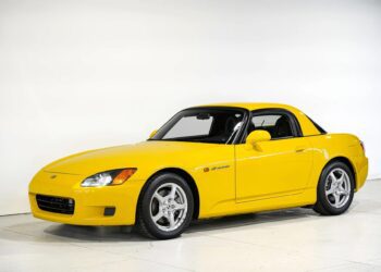 A yellow Honda S2000 sports car with a hardtop roof, one of the best 2000s cars, is parked indoors against a plain white background.
