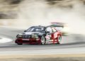A black and red BMW race car speeds around the track at Monterey Car Week 2025, kicking up dust, with another car blurred in the background.