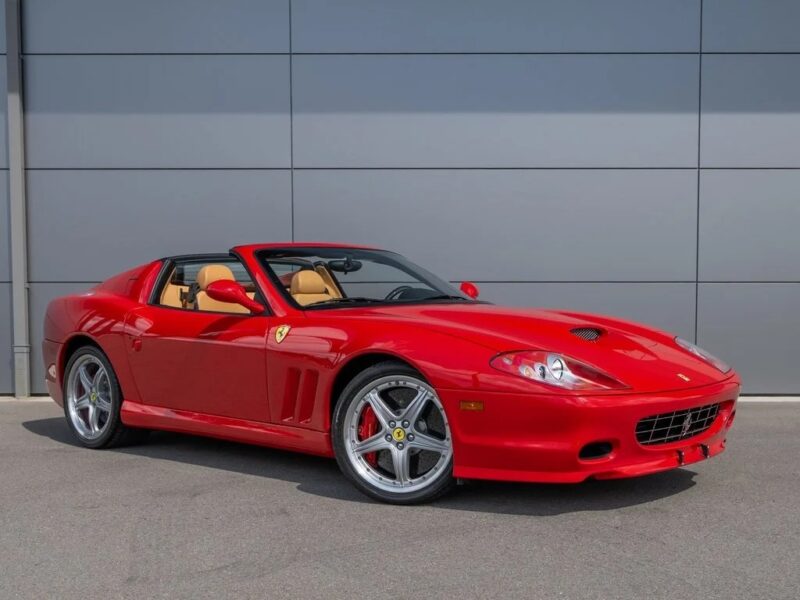 A red Ferrari sports car with tan interior, one of the best 2000s cars, is parked on asphalt in front of a gray metal panel wall.