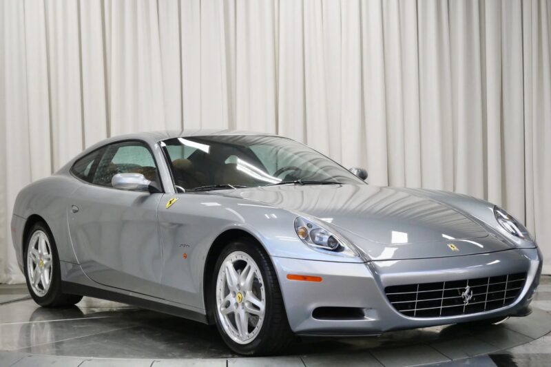 A silver Ferrari coupe, one of the best 2000s cars, is parked indoors on a tiled floor with a white curtain backdrop.