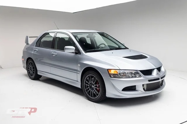 A silver Mitsubishi Lancer Evolution sedan, one of the best 2000s cars, is parked in a white studio, viewed from the front left angle and featuring a hood vent and rear spoiler.
