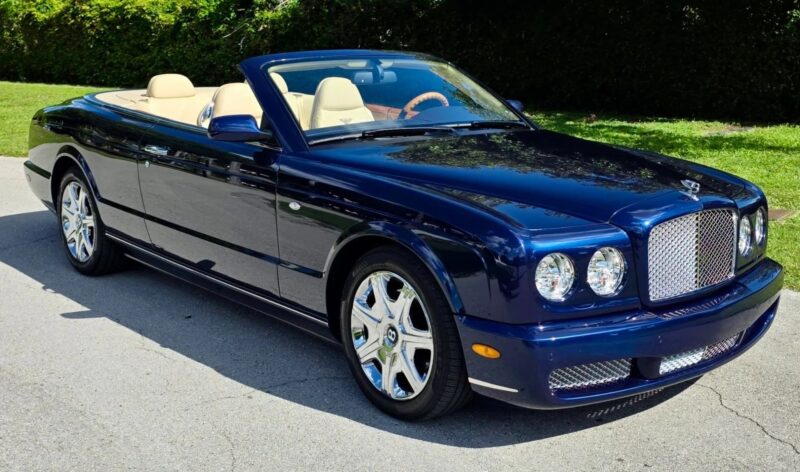 A dark blue luxury convertible car with beige leather interior, one of the best 2000s cars, is parked on a paved road next to green grass and hedges.