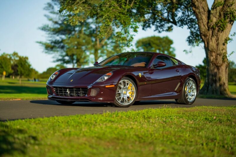 A maroon Ferrari sports car with one of the best manual transmissions is parked on a paved road next to a grassy area with trees in the background on a sunny day.