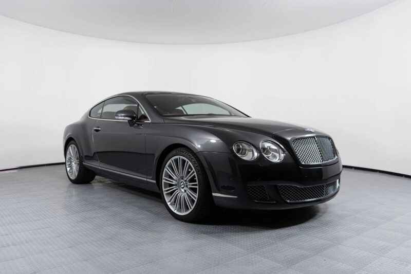 A black Bentley Continental GT coupe, one of the best 2000s cars, is parked in a white-walled showroom, viewed from the front left side.