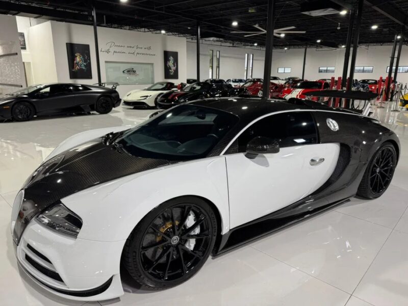 A white and black Bugatti exotic car with a carbon fiber hood is parked indoors among other luxury vehicles in a showroom.
