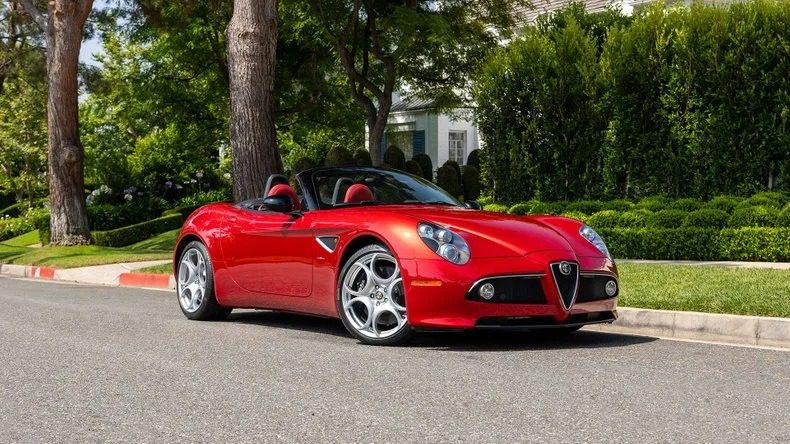 A red luxury convertible sports car is parked on a suburban street, with greenery and a house visible in the background.
