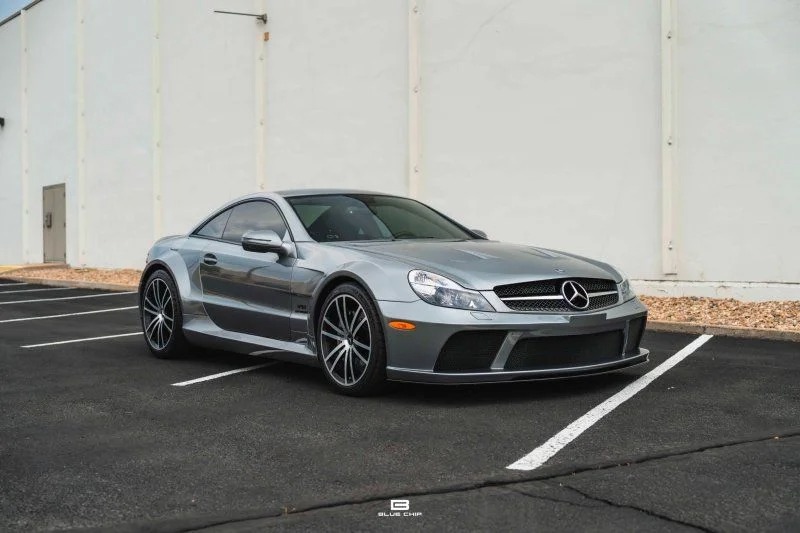 A silver Mercedes-Benz sports car, one of the best 2000s cars, is parked in an empty lot in front of a plain beige wall, photographed from the front left side.