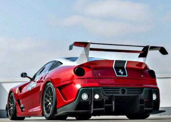 A striking Ferrari 599XX EVO with black accents and a large rear wing is parked outdoors under a partly cloudy sky, awaiting its debut at the Monterey auction.