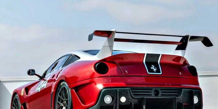 A striking Ferrari 599XX EVO with black accents and a large rear wing is parked outdoors under a partly cloudy sky, awaiting its debut at the Monterey auction.