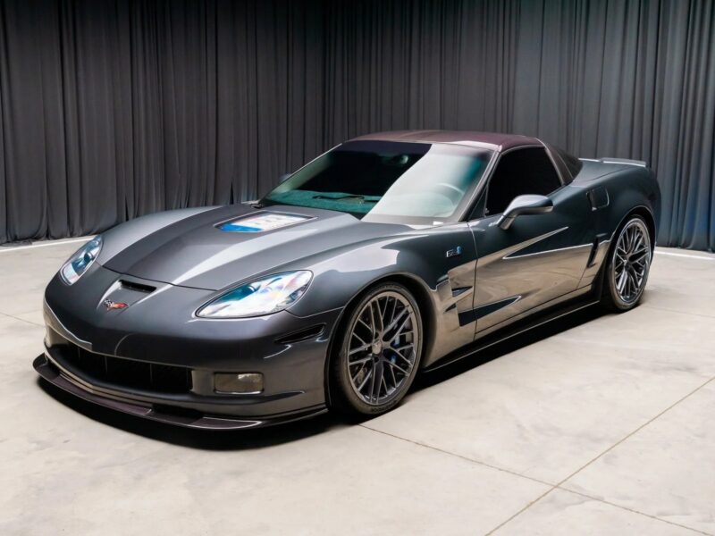 A sleek dark gray Chevrolet Corvette sports car, known among the best manual transmission cars, is parked indoors on a smooth concrete floor, with dark curtains in the background.
