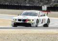 A white BMW race car with the number 55 on the side takes a corner at Monterey Car Week, featuring sponsor decals and a red rear wing.