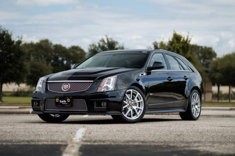 A black Cadillac CTS-V Wagon, one of the finest luxury performance wagons, is parked in an empty lot on a cloudy day, with trees and a building in the background.