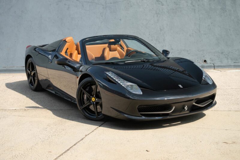 A black Ferrari 458 convertible with tan leather interior is parked on a light-colored concrete surface in front of a gray wall, showcasing why it’s considered one of the best Ferrari models among cars for sale.