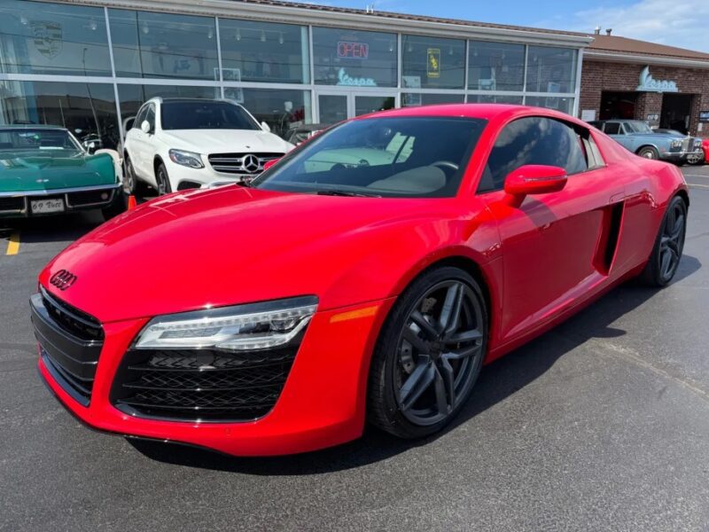 A red Audi R8 exotic car is parked on asphalt in front of a car dealership, with other vehicles visible in the background.