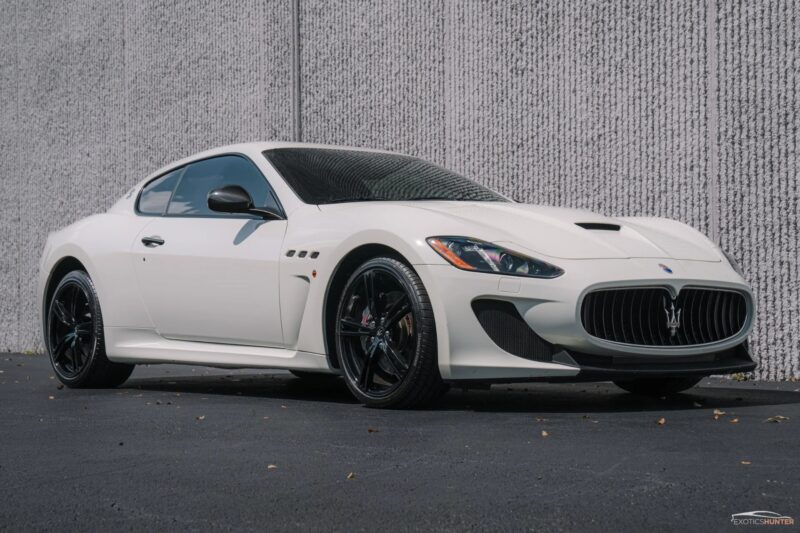 A white Maserati GranTurismo coupe, a true icon of Italian luxury, is parked on a blacktop in front of a textured gray wall.