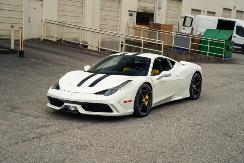 A white Ferrari 458 with black racing stripes is parked on a paved surface in an industrial area near loading docks and a green dumpster, standing out as one of the best Ferrari cars for sale.