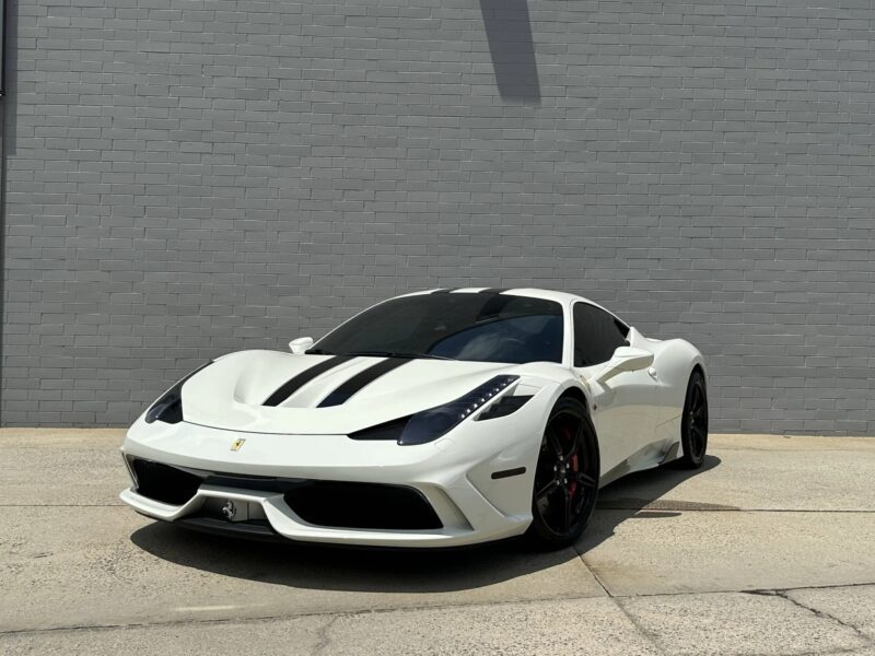 A white Ferrari 458 sports car with black racing stripes is parked on a paved surface in front of a gray brick wall, showcasing why it’s considered one of the best Ferrari models among cars for sale.