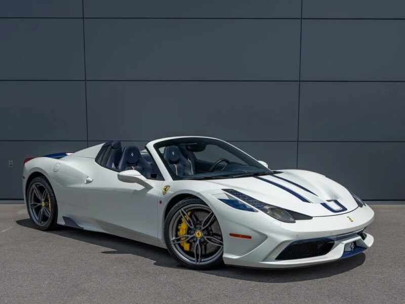 A white Ferrari 458 convertible with blue racing stripes is parked on a paved surface in front of a gray paneled wall, showcasing why it’s considered one of the best Ferrari cars for sale.